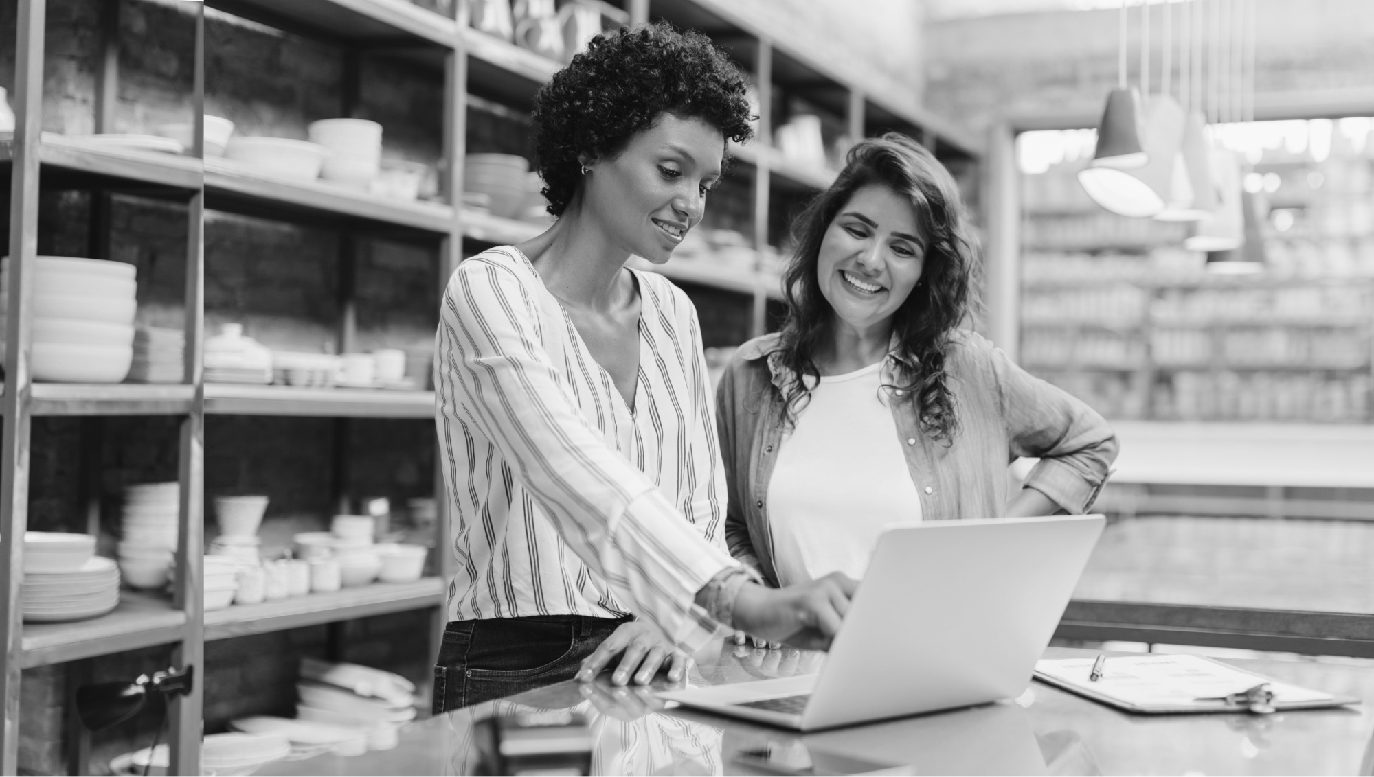 Two women smiling and looking at a laptop together in a creative space BW